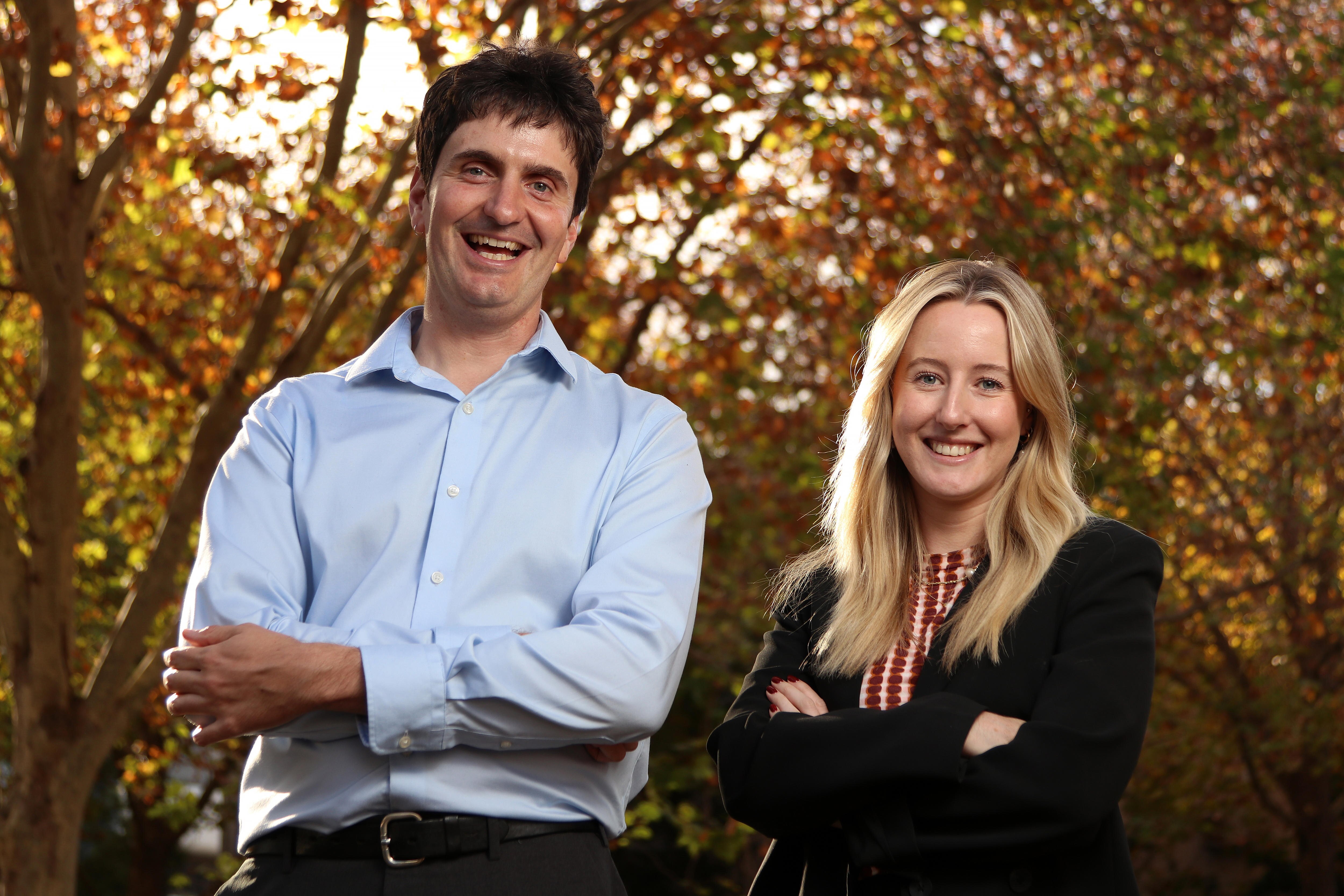 Two people stand beneath a row of trees displaying autumn leaves
