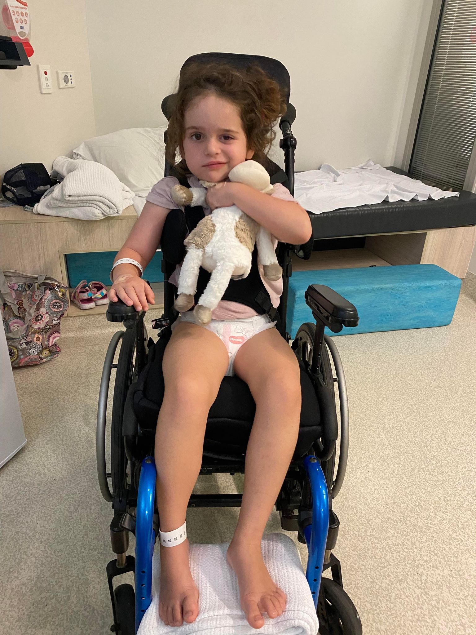 Little girl, Arlo, brown hair, brown eyes, sitting in a wheelchair hugging a toy, hospital room behind.