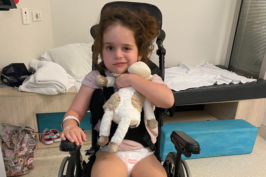 Petite fille, Arlo, cheveux bruns, yeux bruns, assise dans un fauteuil roulant serrant un jouet, chambre d'hôpital derrière.