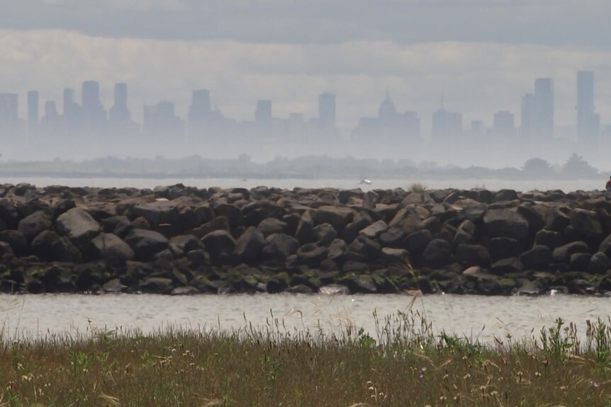 A row of rocks sit in front of the hazy Melbourne CBD skyline, viewed from the bay at Point Cook.