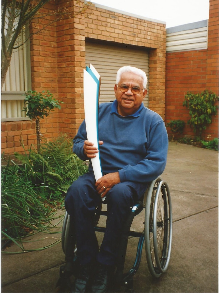 An older Indigenous Australian man with white hair holds a Paralympic torch in his driveway. 