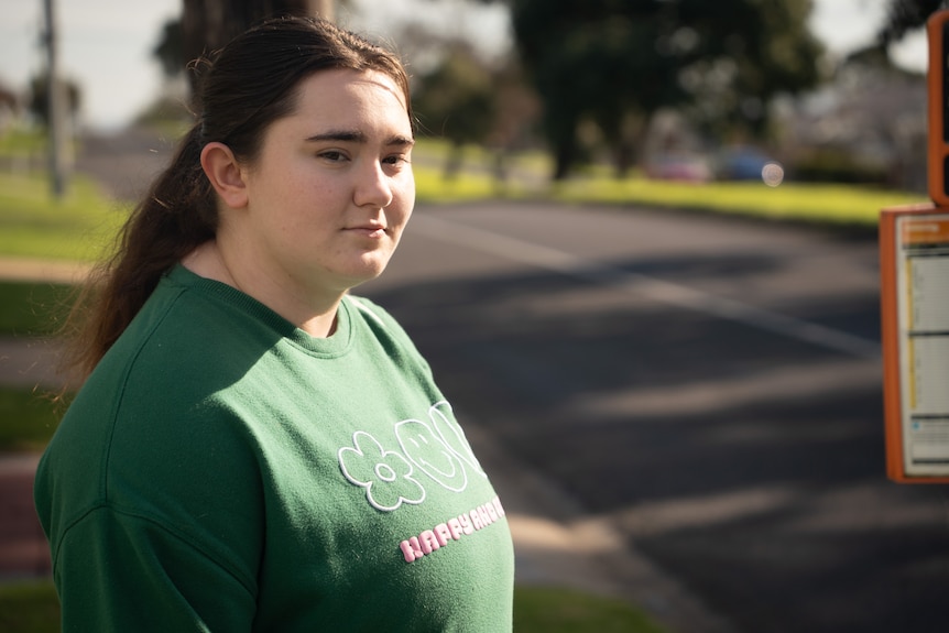 Teenage girl in a green jumper and dark brown hair waits for a bus on an outer Geelong street