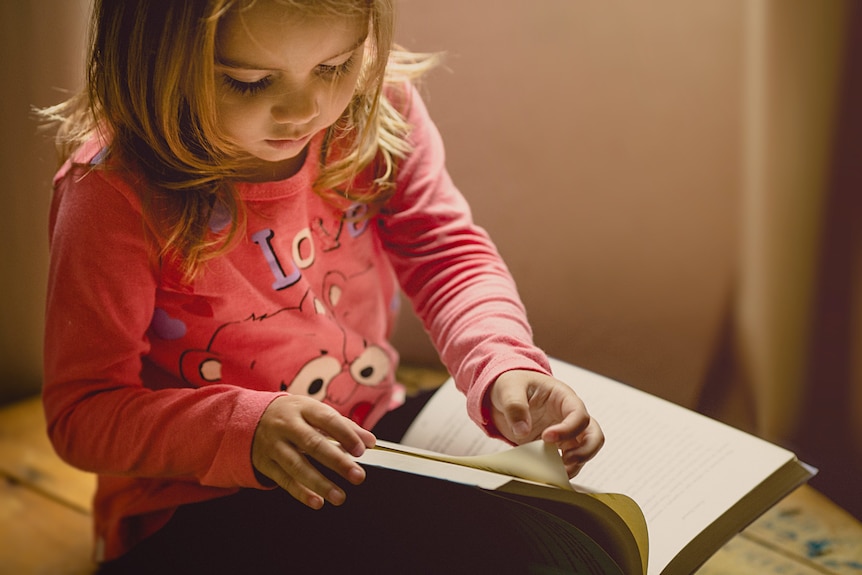 A young girl dressed in a pink top looks down at a book to turn the page.