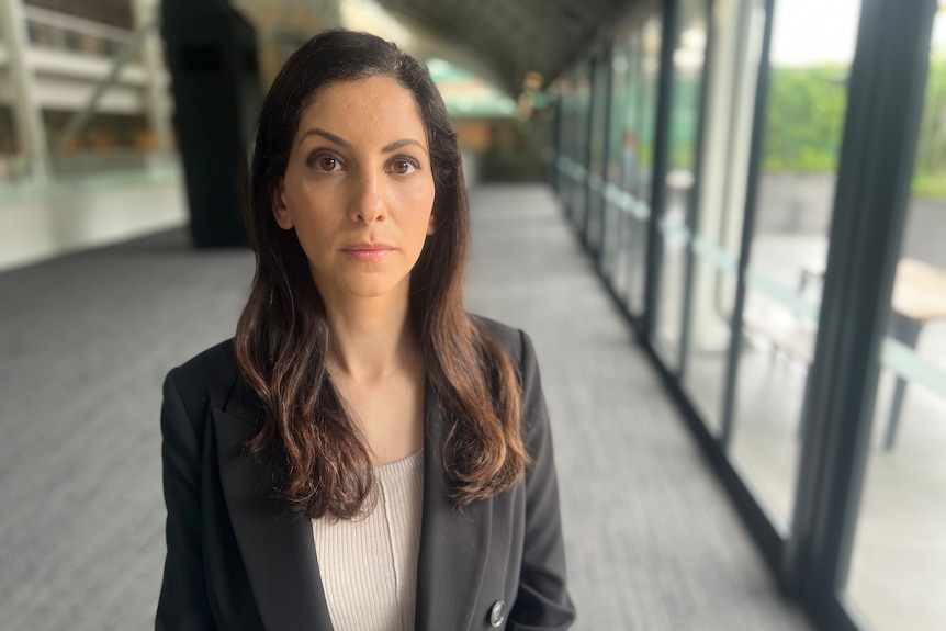 A woman with long dark hair wearing a business jacket poses for a photo inside a modern building.