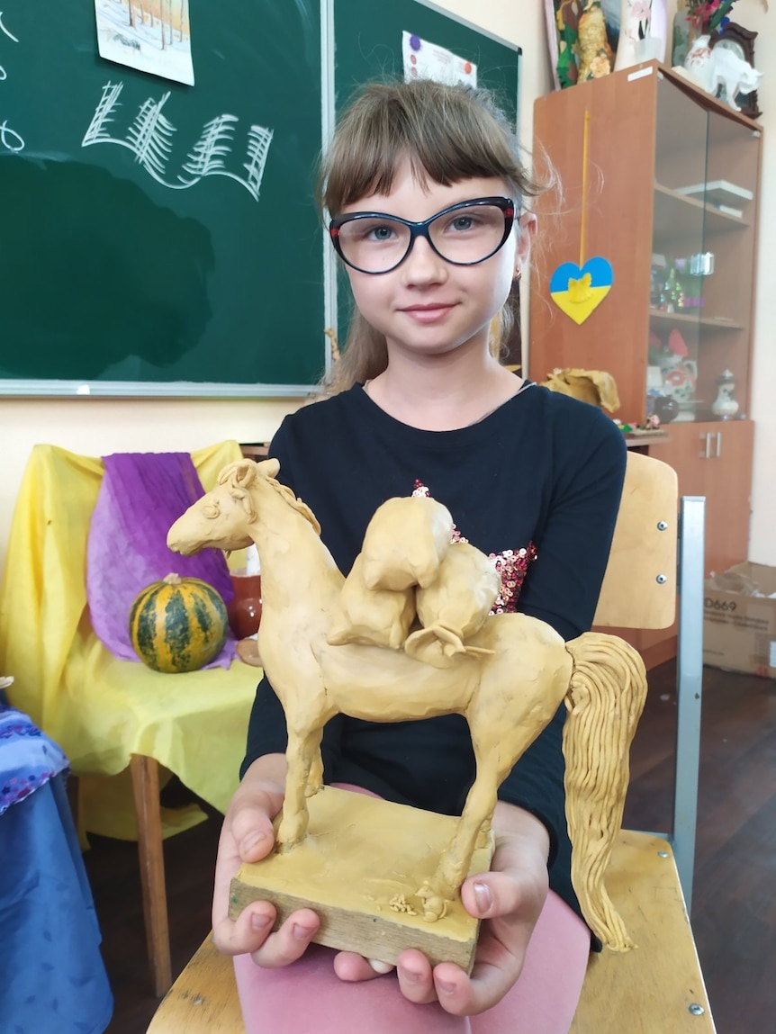 A little girl wearing glasses holds a model of a horse up to the camera, while standing in a classroom