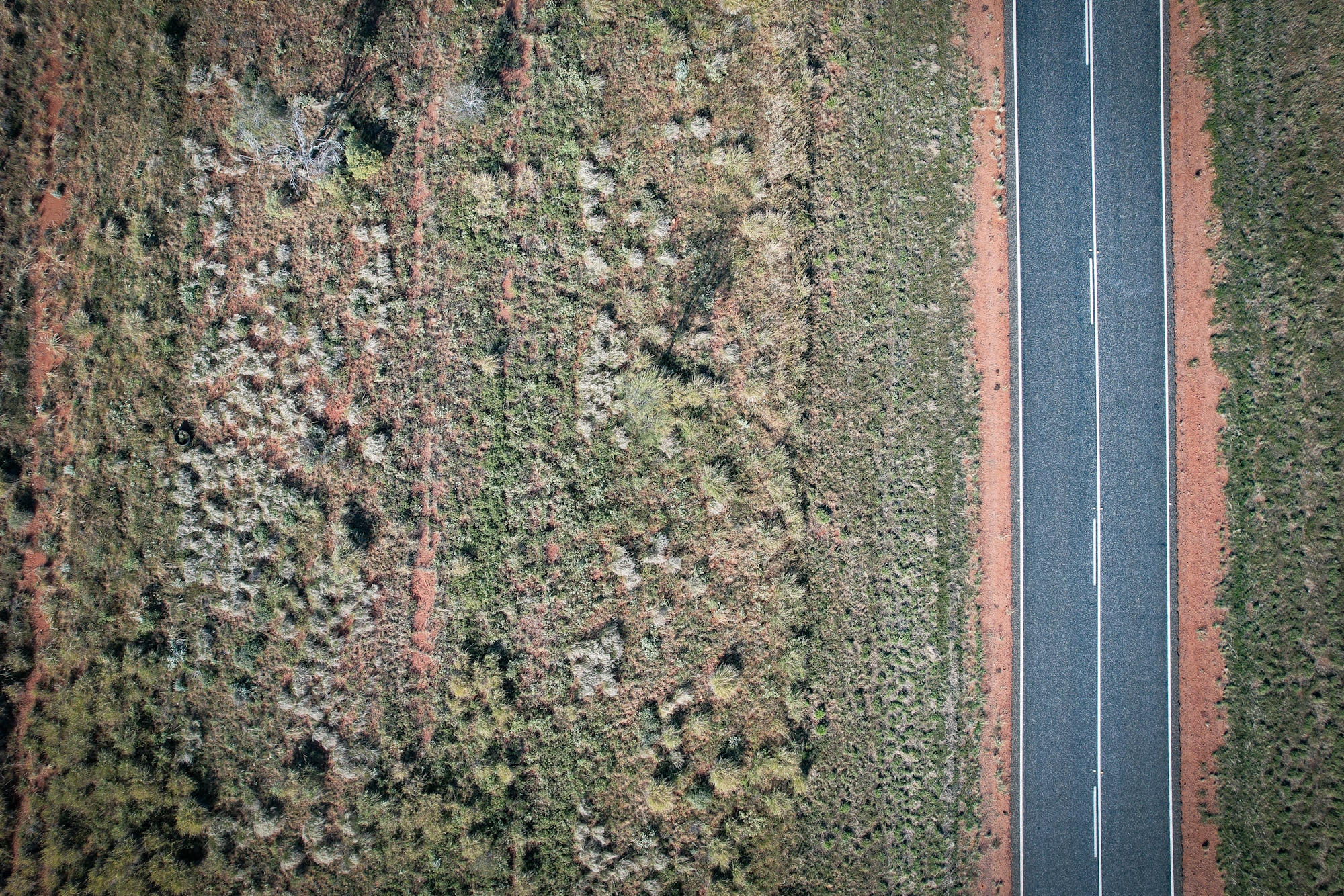 A birds eye view over a remote outback highway, with edges of the black bitumen tapering off into red dirt and green scrubland.