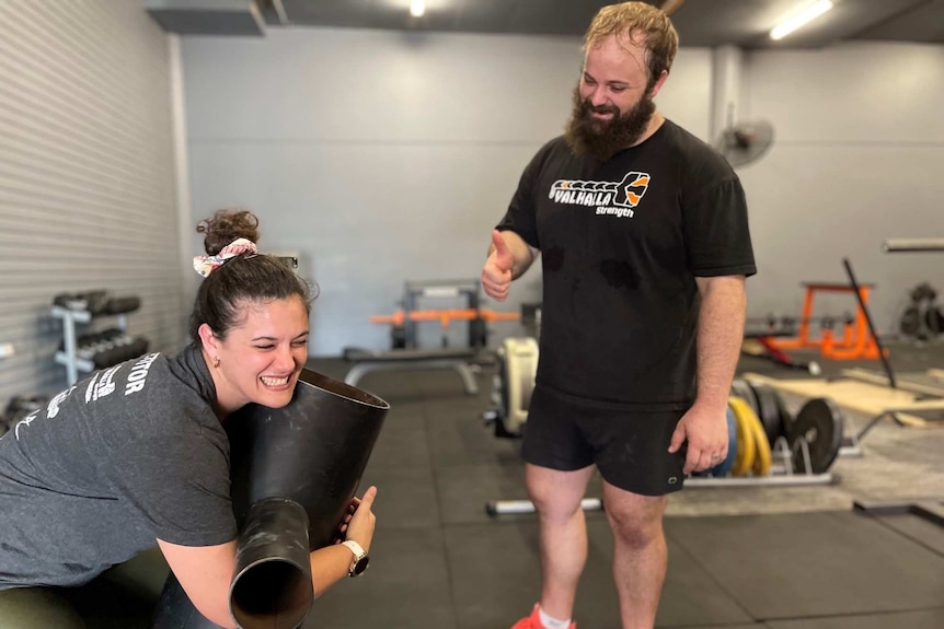 A woman strains as she tries to lift a weight as her coach looks on.