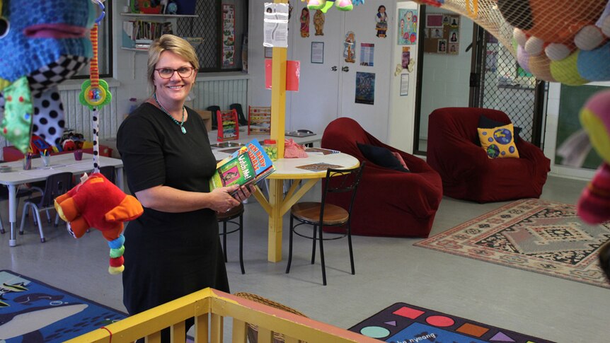 A woman holding children's books stands in a child care centre
