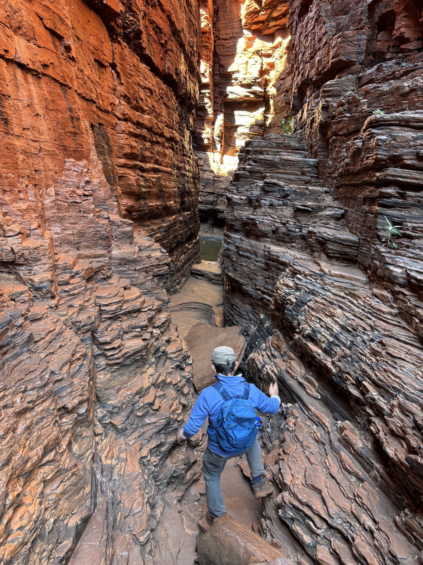 Man faces away from camera wearing a blue long sleeved shirt and backpack, climbing down a browny red rock gorge