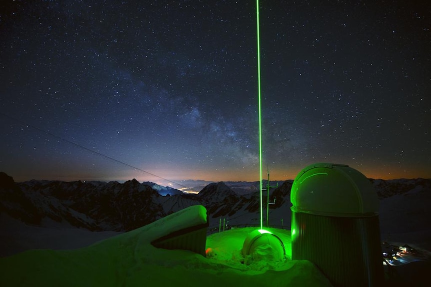 An eerie green laser beam is fired directly up into the night sky from the rooftop