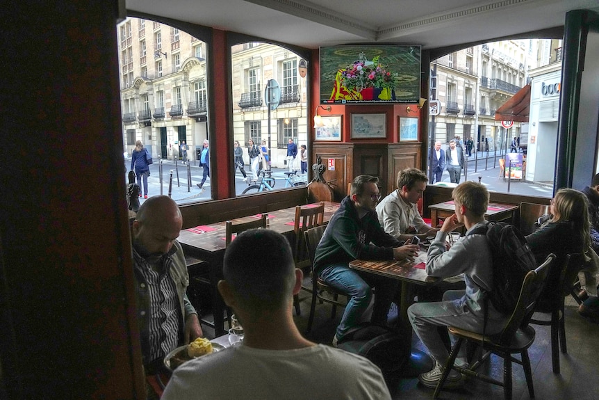 The Queen's funeral plays in a pub as a table of people watch on and people pass by the windows. 