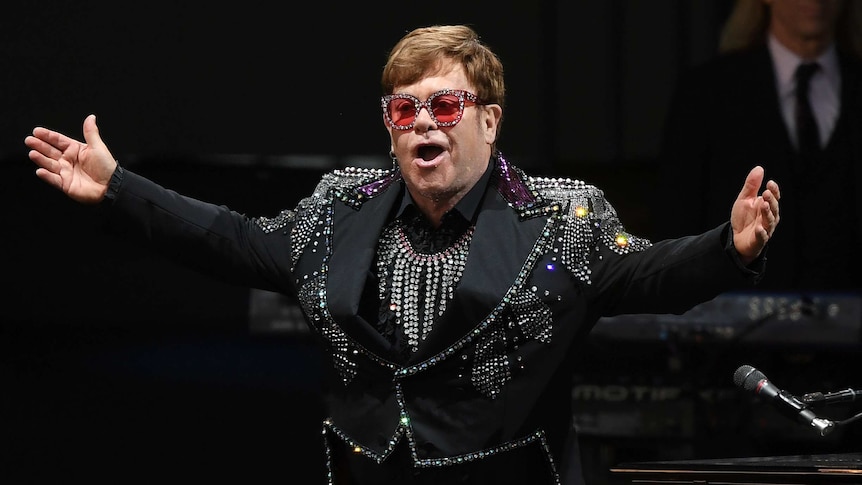 Elton John dressed in a glittery blazer and red sunglasses with his arms out on stage