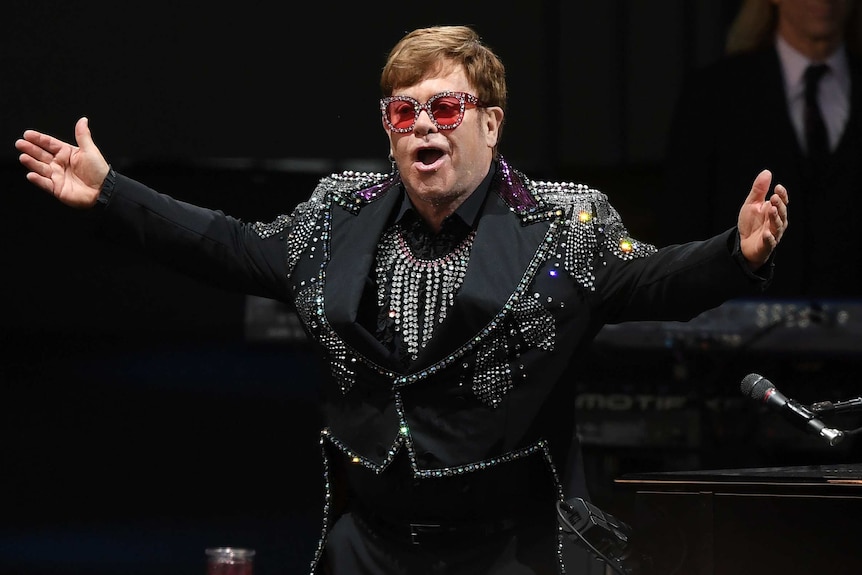 A man dressed in a glittery blazer and red sunglasses with his arms out on stage