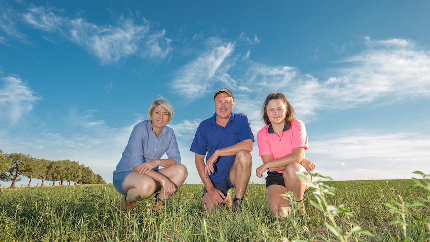 A woman, man and their daughter crouch in a green field of pasture 