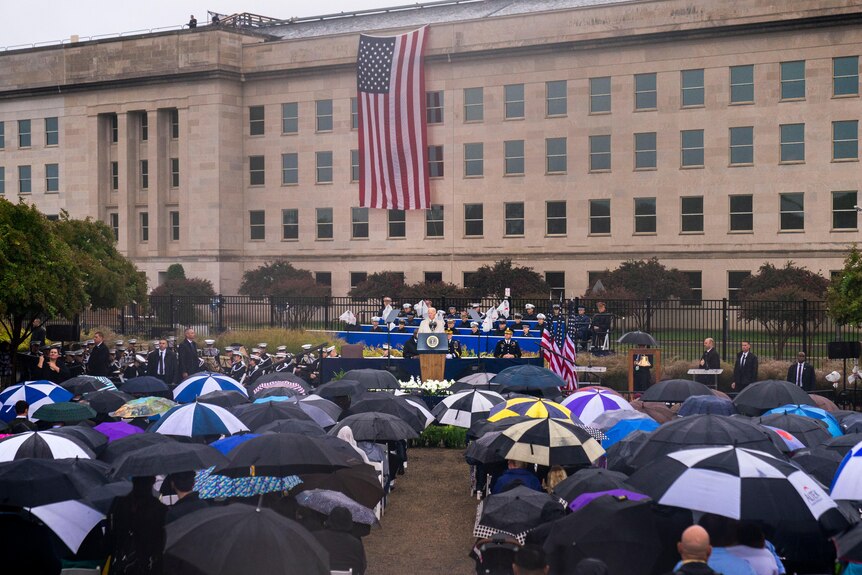 An American flag is draped over a sandy coloured building as a crowd listens to talk on rainy day