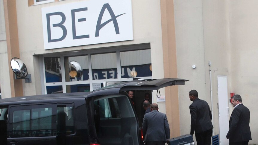 A black van is parked with its boot open as men in dark suits carry a locked grey hard-case into a building marked 'BEA'.