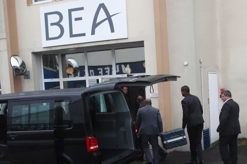 A black van is parked with its boot open as men in dark suits carry a locked grey hard-case into a building marked 'BEA'.