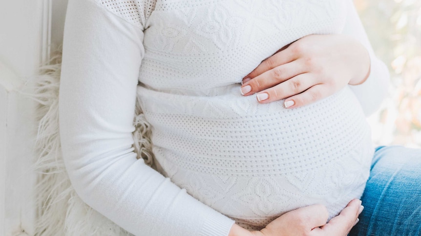 you can see a woman's torso, she is pregnant and holding her tummy she is wearing a white textured jumper and jeans