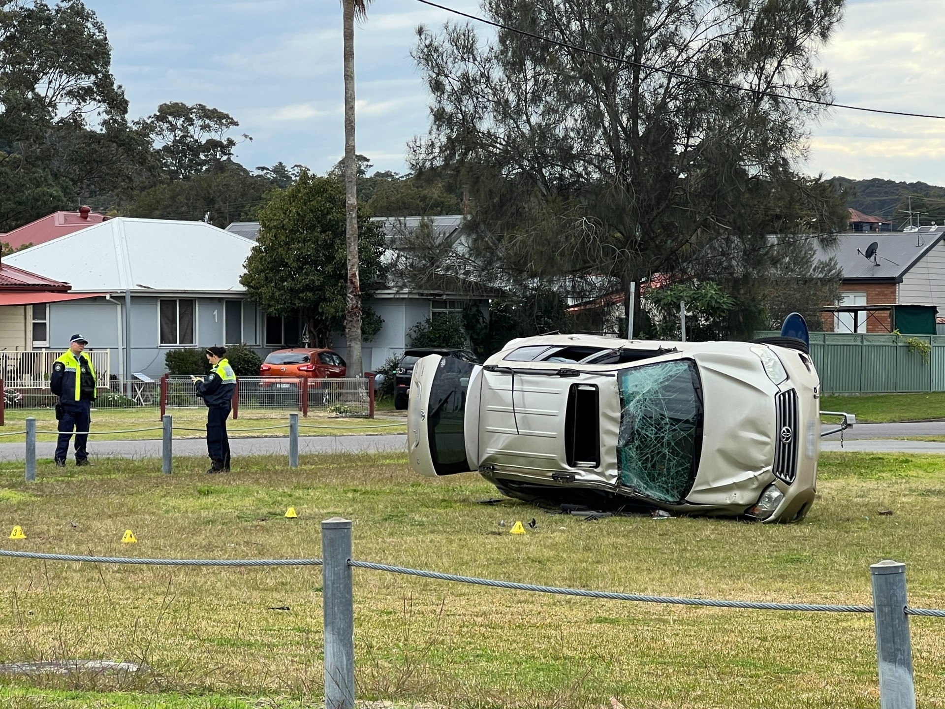 Two police officers stand next to a field. A rolled car with a smashed windscreen lies in the middle.