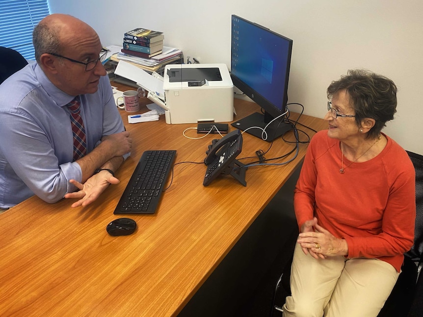 An older male doctor with glasses sits behind a desk with a computer speaking to an older woman patient