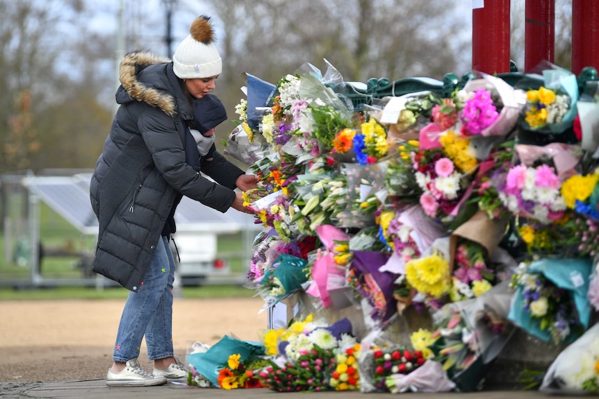 A woman with a baby strapped to her front lays flowers at a large memorial where lot of other bunches of flowers have been left.
