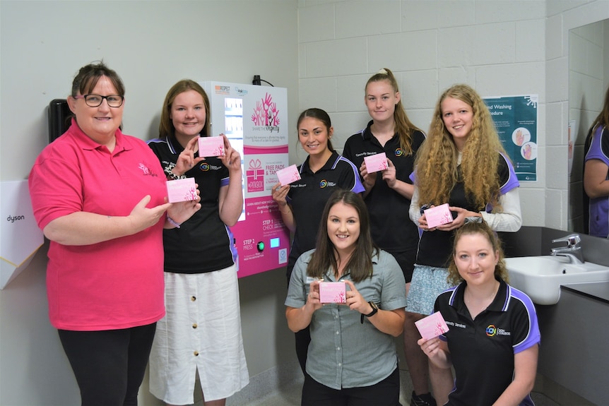 Seven women in a bathroom hold small pink boxes next to a large pink vending machine smiling at the camera.