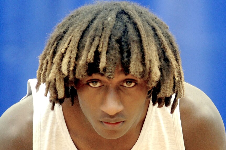 A man with dread hair leans over and stares in a camera
