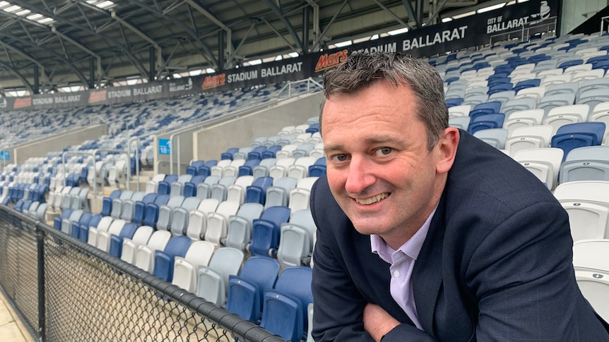 a man wearing a suit leans against the fence of a football stadium with blue and white seats