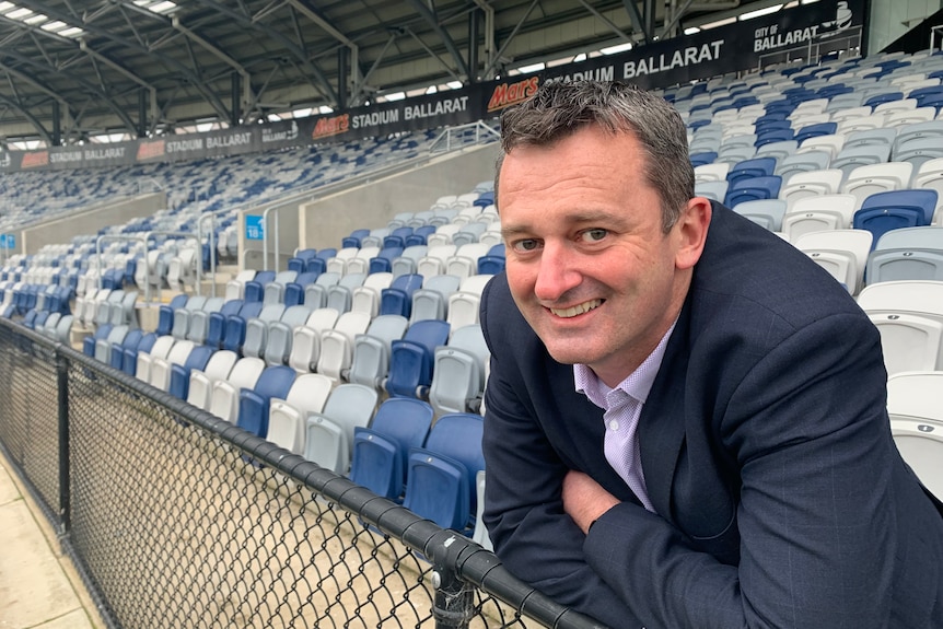 a man wearing a suit leans against the fence of a football stadium with blue and white seats