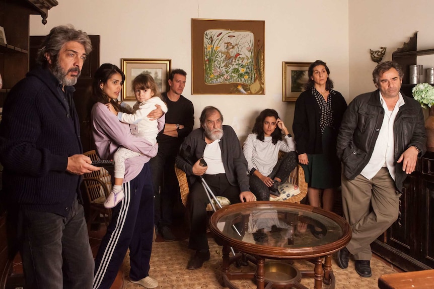 The cast dressed in casual garb stand in a line (two seated on a couch) against the background of an old fashioned living room.
