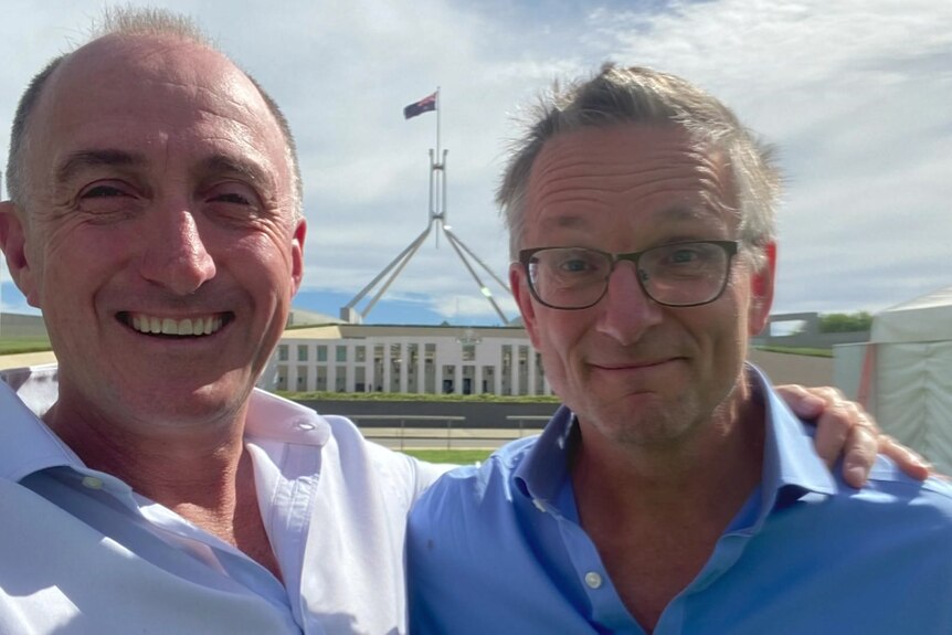 Two men stand in front of parliament house smiling while taking a photo. 