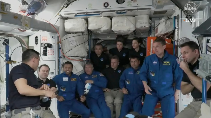 Four new astronauts dressed in blue jumpsuits are greeted by current members dressed in black polos on the ISS.