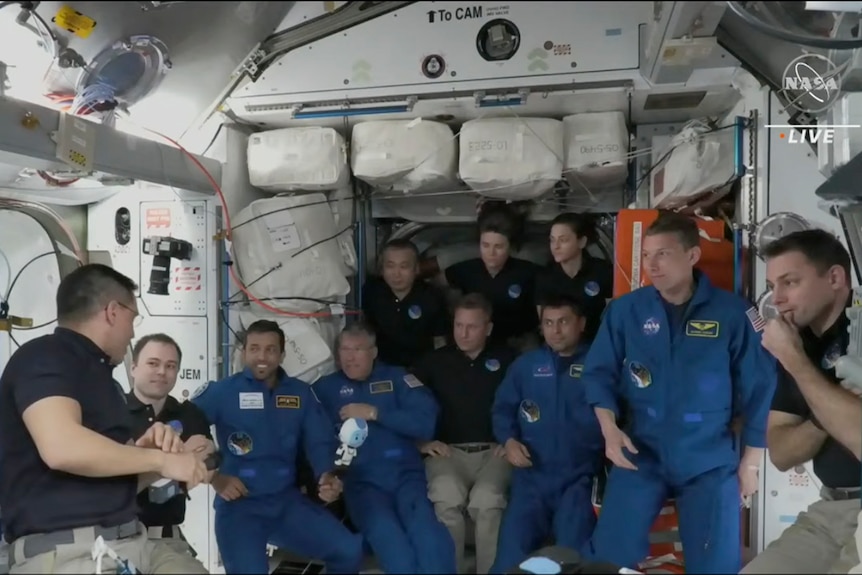 Four new astronauts dressed in blue jumpsuits are greeted by current members dressed in black polos on the ISS.