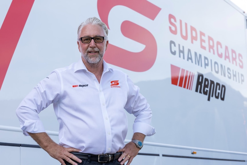 A man stands with hands on hips in front of a Supercars sign.