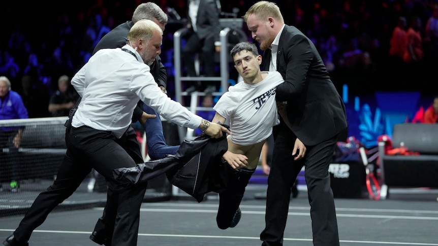 A man in a white t shift is held by three men in suits on a tennis court