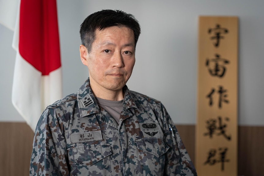 A man in army fatigues stands in front of a Japanese flag