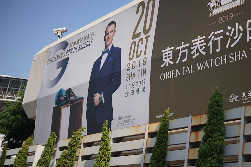 A photo of Zac Purton on a billboard. He’s wearing a navy suit with a bow tie and a watch.