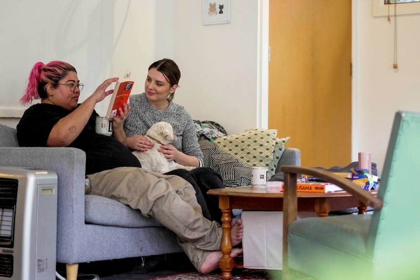 Two people sit on a couch with a small white dog