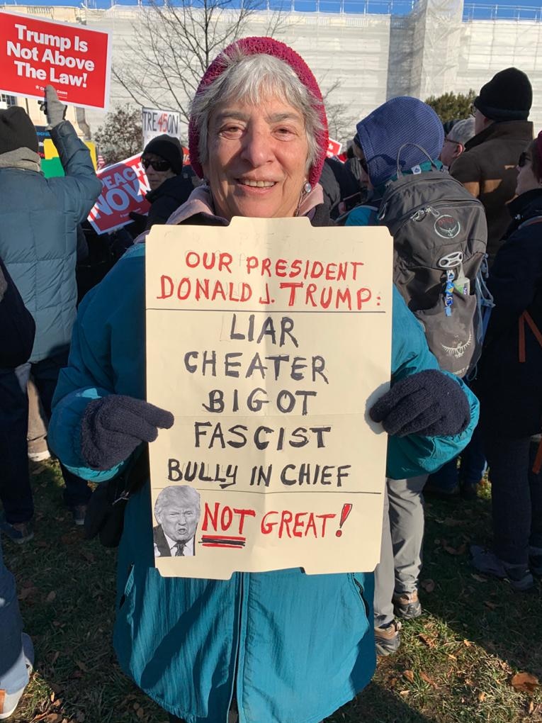 A woman holding a sign reading 'Donald Trump - liar, cheater, bigot, fascist'