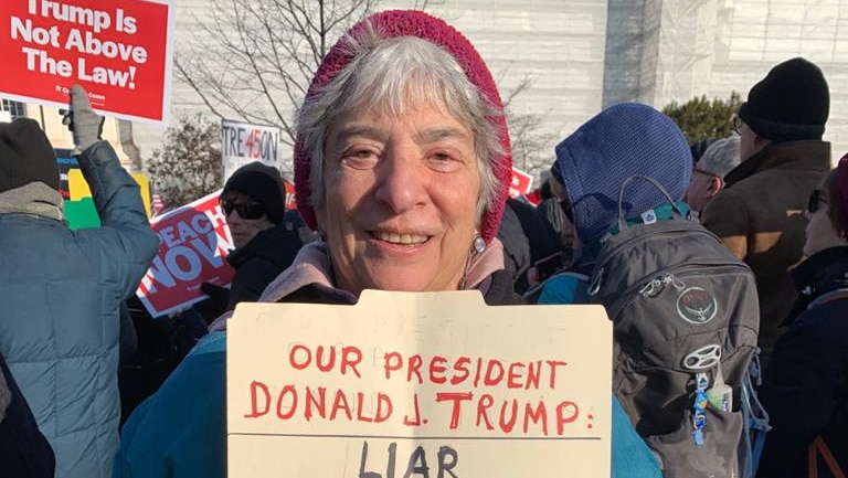 A woman holding a sign reading 'Donald Trump - liar, cheater, bigot, fascist'