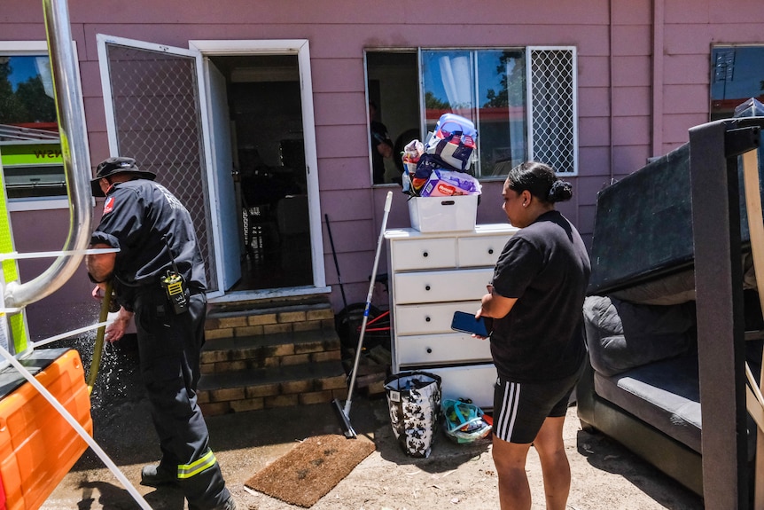 A woman in dark shorts and T-shirt watches as a man uses a high-pressure hose to clean muddy surfaces