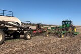 A green tractor pulling a disc seeder in a brown hilly paddock planting wheat seed.