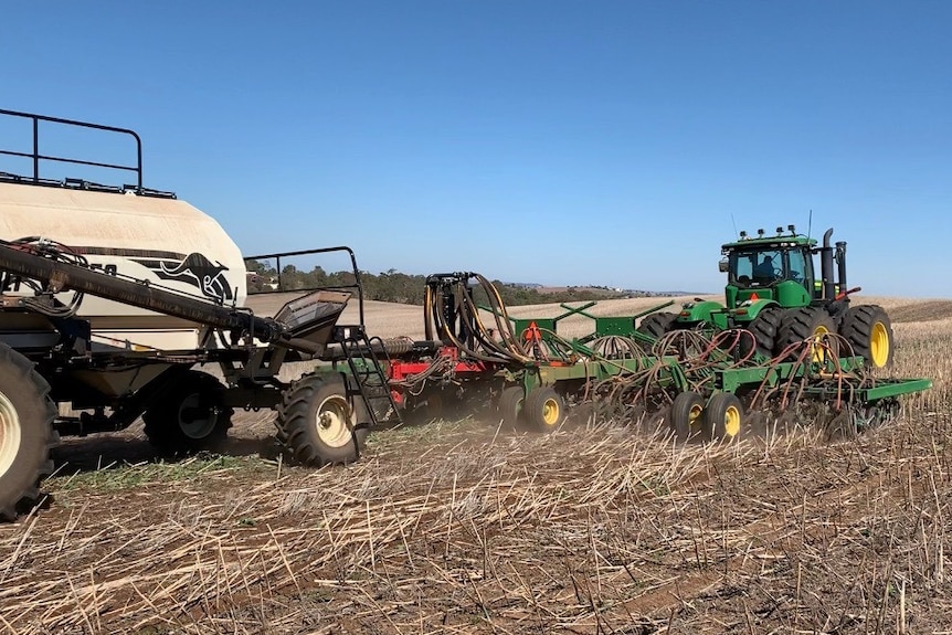 A green tractor pulling a disc seeder in a brown hilly paddock planting wheat seed.
