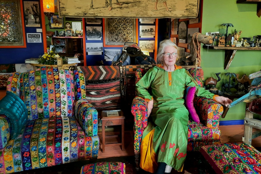 Woman in green dress sits on colourful chair inside room full of pictures, rugs and artefacts.