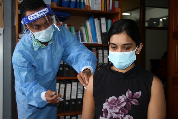 Woman with face mask and black top is vaccinated by worker in mask and medical scrubs.