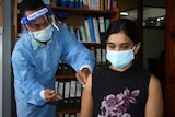 Health worker in blue suit and face mask jabs woman in black top with injection.