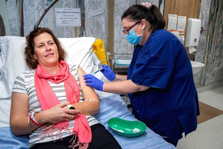 A woman lying on a hospital bed being injected by a nurse wearing a mask