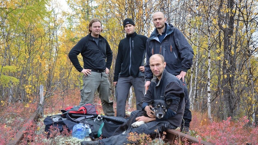 Four men stand in an autumn forest with a pile of survival and technical gear in front of them.