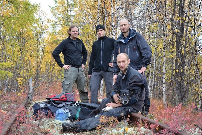 Four men stand in an autumn forest with a pile of survival and technical gear in front of them.