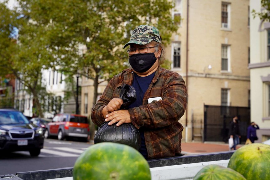 A woman wearing a cap and a black mask holds a black bag on a table with watermelons on it.